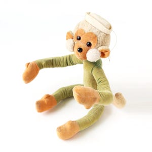 May include: A green plush monkey toy with a white hat, brown eyes, and orange feet. The monkey is sitting with its arms outstretched.
