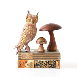 May include: A ceramic owl figurine with a speckled brown and yellow glaze, a wooden mushroom figurine, and a larger wooden mushroom with a brown stem, all arranged on top of two vintage books. The books are titled "Easy" and "Practical Recipes".