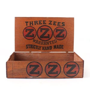 May include: A rectangular wooden box with an open lid. The lid features the text THREE ZEES, three red circles with black Zs, and the words WARRANTED STRICTLY HAND MADE. The front panel displays three red circles with black Zs.