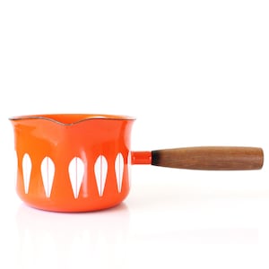 Puede incluir: Cacerola de esmalte naranja con mango de madera. La olla presenta un patrón de gotas blancas alrededor de la base. El mango es de madera y está unido al lateral de la olla. La cacerola está diseñada para cocinar.