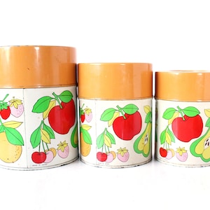 May include: A set of three cylindrical metal canisters with orange lids and white bodies. The canisters feature a colorful graphic print of various fruits including red apples, yellow pears, cherries, and strawberries, with the text Made in Japan.