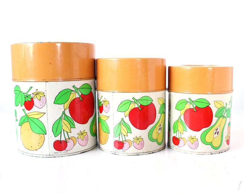 May include: A set of three cylindrical metal canisters with orange lids and white bodies. The canisters feature a colorful graphic print of various fruits including red apples, yellow pears, cherries, and strawberries, with the text Made in Japan.