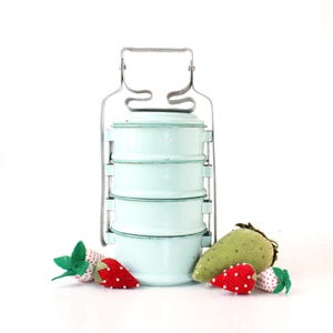 May include: A vintage, light blue, metal tiered lunch box with a silver handle. The lunch box is stacked with four containers and is surrounded by felt strawberries.