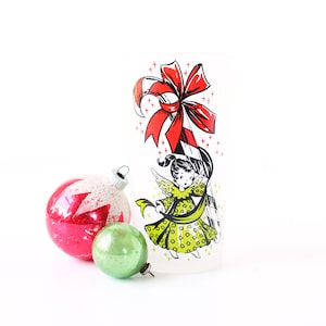 May include: Vintage Christmas ornaments and a glass with a festive angel design. The ornaments include a red and white glittered ball, and a small green ball. The glass features a red bow and a cartoon angel in a green dress.