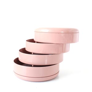 May include: A stack of four pink, circular storage trays. The trays are arranged in a spiral, with each tray slightly offset from the one below. The top tray has a lid. The trays are made of a smooth, solid material and are set against a white background.