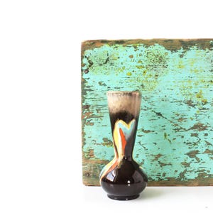 Puede incluir: Un jarrón de cerámica vintage con base marrón oscuro y cuello acampanado. El jarrón presenta un diseño colorido con detalles en azul, naranja y amarillo. El fondo incluye un panel de madera turquesa desgastado.