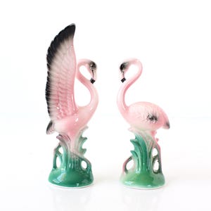 Puede incluir: Dos figuras de flamencos de cerámica. Uno tiene un ala negra y rosa extendida. El otro flamenco está de pie. Ambos son rosas con detalles negros y están sobre bases verdes.