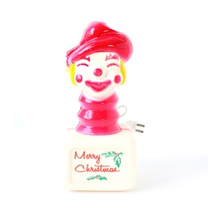 Könnte beinhalten: Ein Vintage-Weihnachts-Nachtlicht in Form eines lächelnden Clowns mit rotem Hut und Outfit. Das Gesicht des Clowns ist weiß mit roten Akzenten. Die Basis ist ein weißes Rechteck mit dem Schriftzug "Merry Christmas".