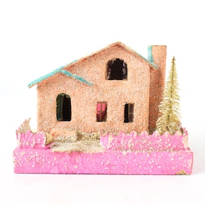 Puede incluir: Una casa en miniatura vintage con una base rosa y un exterior texturizado color melocotón. El tejado es turquesa y un pequeño árbol dorado se encuentra junto a la chimenea. La casa tiene ventanas abiertas y una valla rosa decorativa.