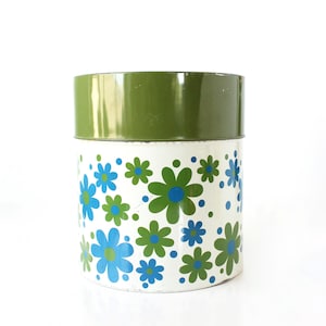 May include: A cylindrical metal canister featuring a solid olive green lid and a white base decorated with a repeating pattern of blue and olive green daisy-like flowers interspersed with small blue and green dots across the entire surface.