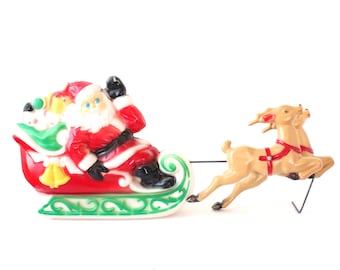 May include: A vintage Christmas decoration featuring Santa Claus in a red sleigh pulled by two reindeer. Santa is wearing a red suit with a black belt and boots. The sleigh is red with green and white accents. The reindeer are tan with red harnesses.