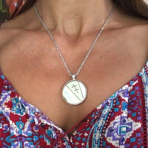 May include: Silver-toned necklace with a round pendant. The pendant features a light green background with two parallel lines and a series of blue and white shapes. The necklace is displayed against a patterned red, blue, and purple top.