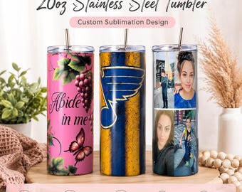 May include: Three 20oz stainless steel tumblers with lids and straws. The left tumbler is pink with grapes, a butterfly, and the text 'Abide in me'. The center tumbler has a blue and gold winged logo. The right tumbler features a photo collage.