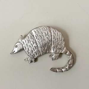 May include: A silver-toned metal brooch shaped like an armadillo. The piece features a textured shell with distinct segmented plates, a pointed snout, small ears, and a long, curved tail. The surface has a matte metallic finish throughout.