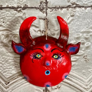 May include: A red, round, devil-faced mask with curved horns, blue accents, and yellow eyes. The mask has a surprised expression with a small, open mouth. A string is attached for hanging.