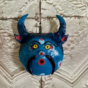 May include: A blue, round, decorative mask with curved horns and a detailed face. The mask features yellow eyes, a red mouth, and black mustache-like shapes. The background is a textured, off-white surface.