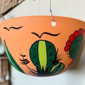 May include: A terracotta-colored hanging planter with a painted design of cacti and birds. The planter has a hole in the top for hanging.