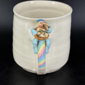 May include: A white ceramic mug with a handle shaped like a light blue creature eating a brown and tan pie. The handle features a pastel rainbow swirl pattern. The mug has a smooth, cylindrical body with subtle horizontal ridges along the exterior.