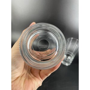 May include: Clear glass tumbler with a ridged design around the base and a smooth rim. The bottom of the glass has the text "WIESER SCHOLL" etched into the glass. Another glass is visible in the background.