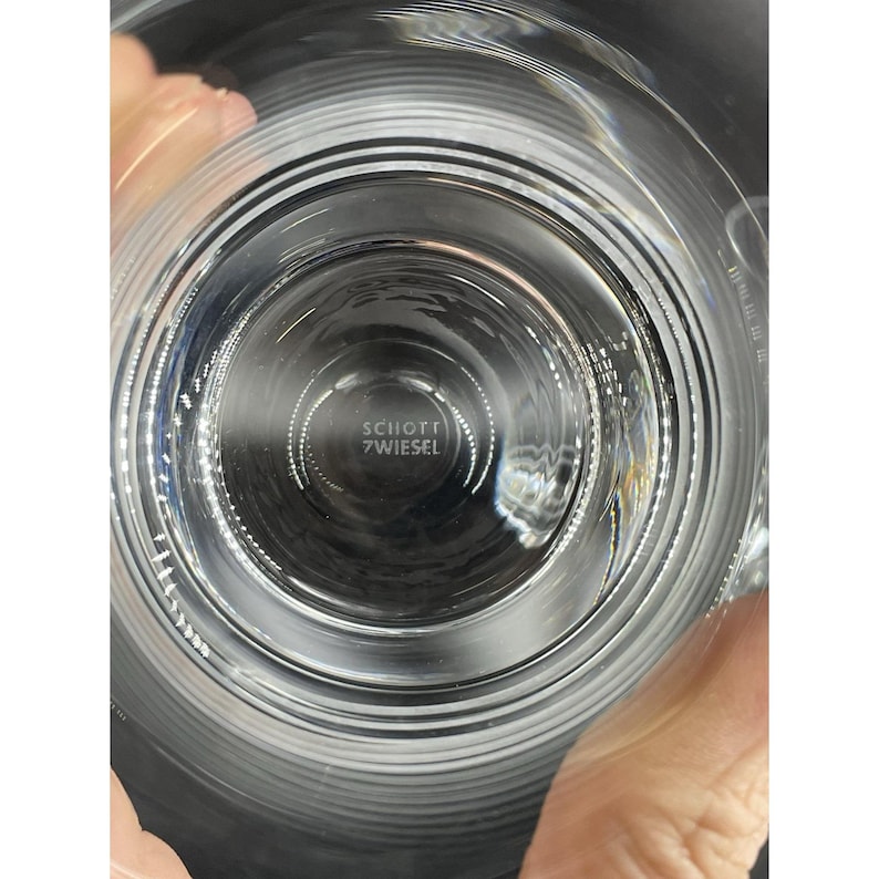 May include: A clear glass with concentric circular ridges. The text "SCHOTT 7 WIESEL" is visible at the bottom of the glass. The glass is held by a person's hands.