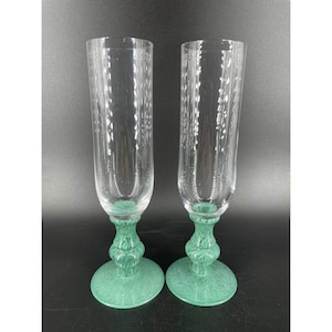 Puede incluir: Dos copas de champán transparentes con base y tallo verdes texturizados. Las copas altas y delgadas tienen un patrón decorativo grabado en el cristal. La base y el tallo verdes añaden un toque de color al elegante diseño.