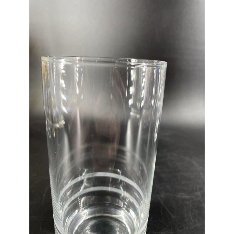 May include: A clear, cylindrical glass with a slightly flared rim. The glass has three horizontal etched lines near the base. The glass is transparent, suitable for beverages. The height is approximately 15 cm.