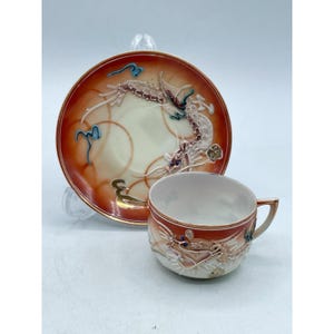May include: A vintage teacup and saucer set. The cup and saucer are decorated with a dragon motif in shades of red, cream, and gold. The saucer has a dragon design and the cup has a matching dragon design. The set is likely made of porcelain.
