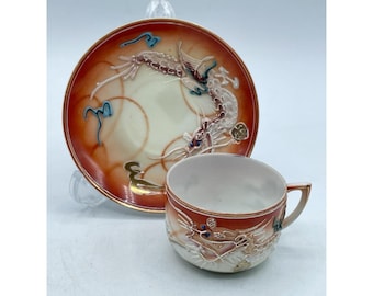 Könnte beinhalten: Ein Vintage-Tee-Set, bestehend aus einer Tasse und einer Untertasse. Die Tasse und die Untertasse sind mit einem Drachenmotiv in Rot-, Creme- und Goldtönen verziert. Die Untertasse hat ein Drachendesign und die Tasse ein passendes Drachendesign.