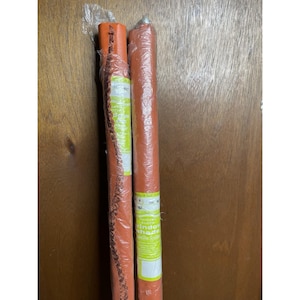 Op de afbeelding: Twee opgerolde oranje stoffen raamjaloezieën, verpakt in doorzichtig plastic. Eén jaloezie heeft een decoratieve rand. De labels op de jaloezieën zijn gedeeltelijk zichtbaar, met de woorden "window shade" erop gedrukt.