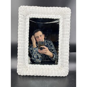May include: White wicker framed mirror with a woven design. The mirror reflects a person taking a selfie.