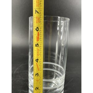 May include: A clear glass tumbler with three white horizontal stripes. A yellow measuring tape is placed next to the glass, showing a height of approximately 17.78 cm.