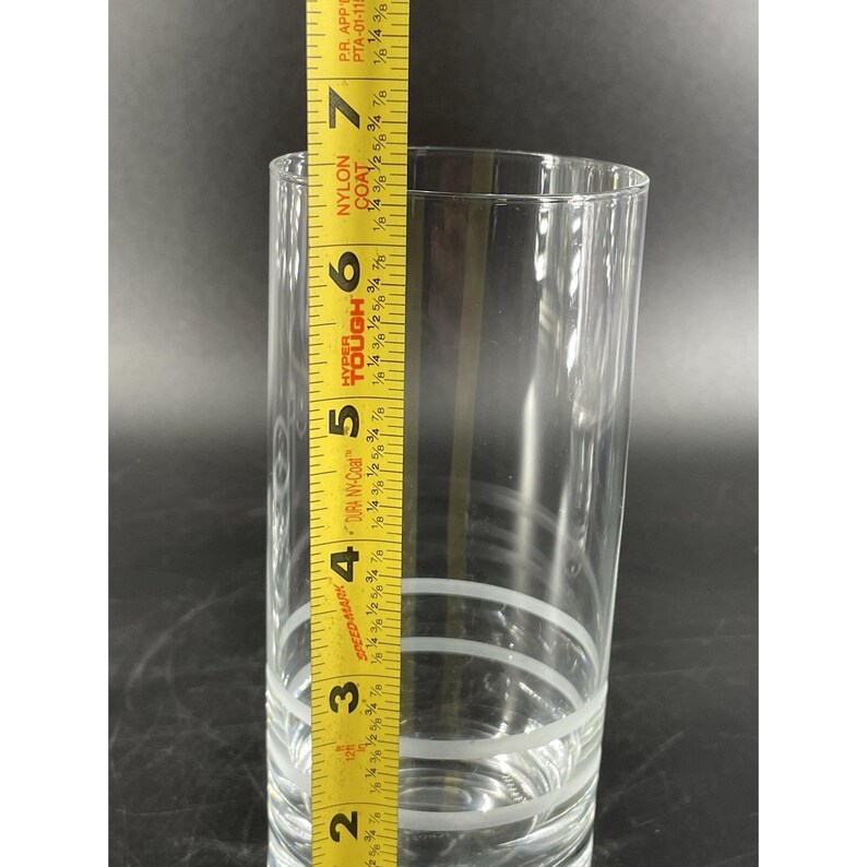 May include: A clear glass tumbler with three white horizontal stripes. A yellow measuring tape is placed next to the glass, showing a height of approximately 17.78 cm.