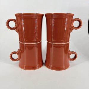 Może przedstawiać: Cztery ceramiczne kubki w kolorze terakoty ułożone w dwie kolumny po dwa. Każdy kubek ma gładkie, błyszczące wykończenie, cylindryczny korpus z lekko rozszerzaną podstawą oraz mały, okrągły uchwyt przymocowany do boku górnej części.