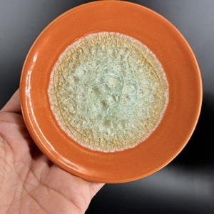 Pode incluir: Um prato redondo de cerâmica com uma borda lisa de cor laranja sólida e um centro texturizado e craterizado num tom verde-menta pálido e translúcido. A área central apresenta um padrão de superfície cristalino e rachado que contrasta.
