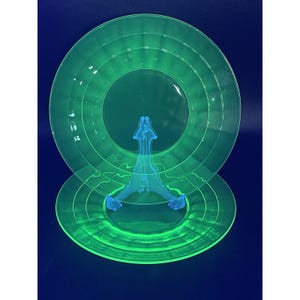 Könnte beinhalten: Zwei grüne Glasplatten mit konzentrischen Ringen, präsentiert auf einem blauen Kunststoffständer. Die Platten haben ein lebendiges, neonartiges Leuchten, vor einem dunkelblauen Hintergrund. Die Platten sind rund und scheinen Vintage zu sein.