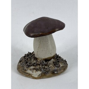 May include: A ceramic mushroom figurine with a brown cap and a white speckled stem. The mushroom is sitting on a base that is textured to look like dirt and grass.