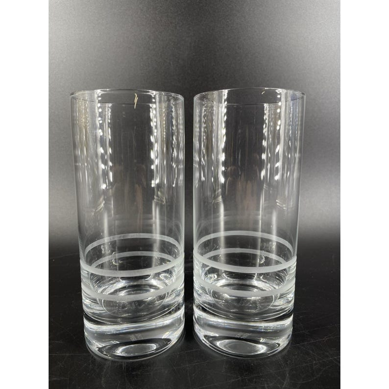May include: A pair of clear, highball glasses. Each glass has a simple design with two etched horizontal bands near the base. These glasses are ideal for serving water, juice, or cocktails. The glasses are approximately 15 cm tall.