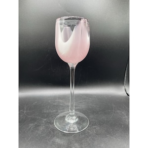 May include: A tall, clear wine glass with a long stem and a round base. The bowl of the glass is a soft pink color with a white, swirling design. The glass is empty and appears to be made of glass.