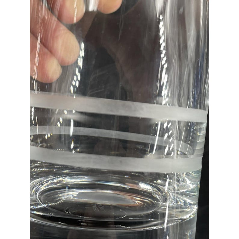 May include: A clear glass with frosted horizontal bands. The bands encircle the glass, creating a decorative pattern. The glass is partially filled with a clear liquid, and the bottom of the glass is visible.
