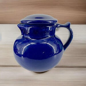 May include: A cobalt blue ceramic pitcher with a matching lid. The pitcher has a rounded body, a curved handle, and a spout for pouring. The glossy finish reflects light, highlighting its smooth surface. A classic kitchenware item.