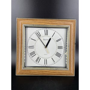 May include: Square Bulova quartz clock with a light brown wooden frame. The clock face is white with black Roman numerals and hands. The clock reads "Bulova Quartz".