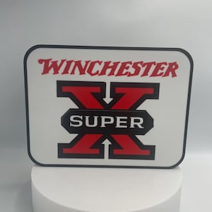 May include: A white square sign with a black border and red and black lettering. The sign says "Winchester" in a stylized font and "Super" in a black rectangle on a red "X" shape.