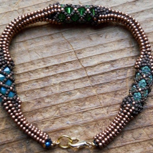 May include: A beaded bracelet with copper-coloured beads and teal and green crystal accents. The bracelet has a gold-coloured clasp and a dark blue bead. The beads are arranged in a spiral pattern.
