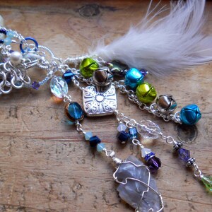 May include: A handcrafted, whimsical charm featuring a small glass bottle, chainmail, and dangling beads in shades of blue, green, and purple. Accented with silver-toned metal charms, bells, and a white feather. A unique, decorative accessory.