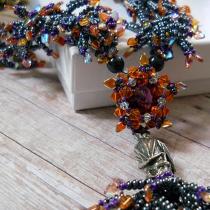 May include: A handmade necklace with a central pendant of orange and purple beads, a silver skull charm, and black beads. The necklace is displayed against a white box and a wooden surface.