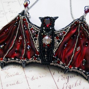 May include: A handcrafted bat pendant necklace. The bat features deep red satin wings, black beaded body, and is adorned with red and clear crystals. The pendant hangs from a silver chain, creating a gothic and elegant accessory.