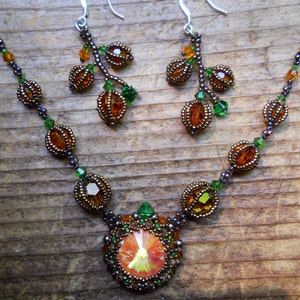 May include: A handmade jewellery set comprising a necklace and earrings. The necklace features a large, orange crystal pendant surrounded by smaller beads. The earrings and necklace incorporate orange, green, and bronze-coloured beads, creating a nature-inspired design.