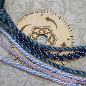 May include: A circular wooden kumihimo disk with numbered markings from 1 to 32, featuring a central hole and decorative etched patterns. Several braided cords are layered over the disk, including strands with small blue and pink beads.