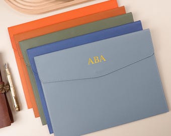 May include: A collection of leather document holders in various colors, including orange, green, blue, and gray. The gray holder has the initials "ABA" in gold. A brown leather journal and a gold pen are also present.