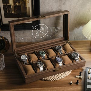 May include: A wooden watch display case with a clear glass top, showcasing ten wristwatches with various band and face designs. The case is open, revealing brown cushions for each watch. The background includes a lamp and a framed picture.